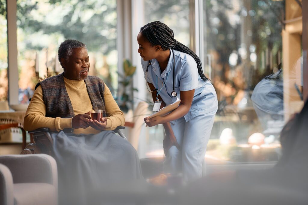 Young black nurse talking to senior man in wheelchair while he is drinking tea in nursing home.