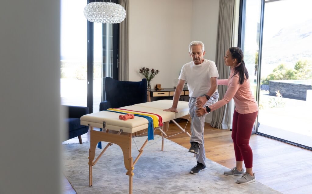 Biracial female physiotherapist assisting caucasian senior man in stretching leg by table at home