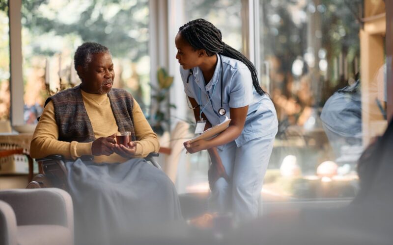 Young black nurse talking to senior man in wheelchair while he is drinking tea in nursing home.
